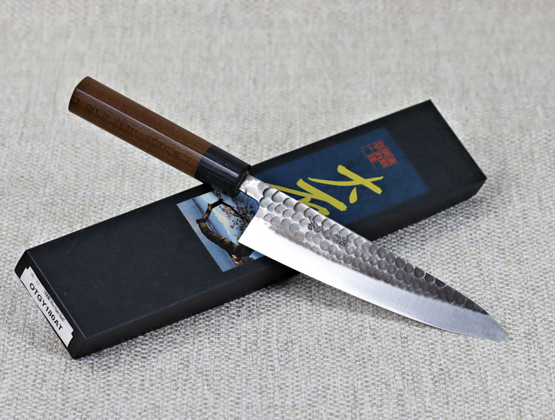 Ohishi Aogami 2 Kurouchi/Tsuchime 180mm Gyuto(Chef) kitchen knife resting at an angle across its packaging