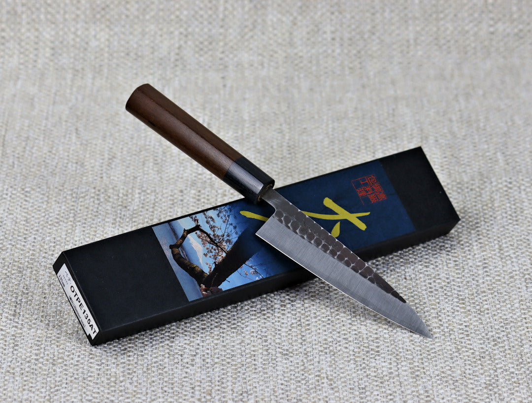 Ohishi Aogami 2 Kurouchi/Tsuchime 135mm Petty (Utility) Kitchen Knife resting at an angle across its packaging