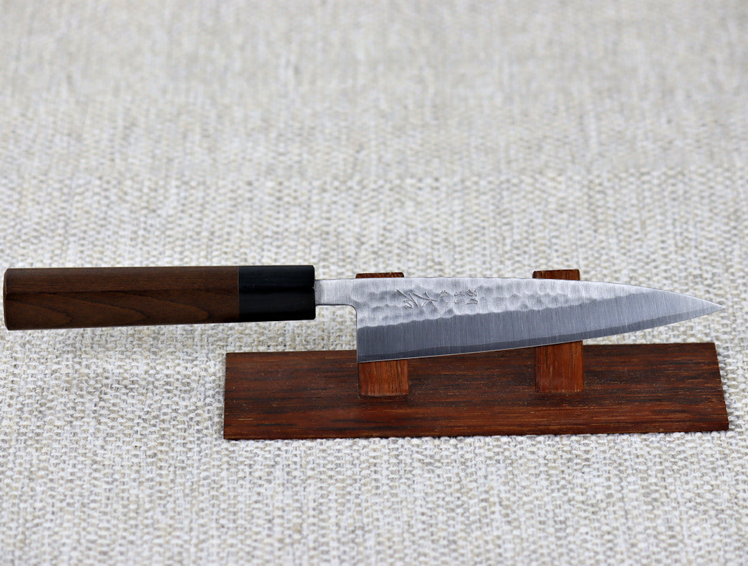 Ohishi SD 135mm Petty Utility Japanese kitchen knife with migaki(polished/tsuchime(hammered) finish and traditional Ebony handle resting on a redwood stand