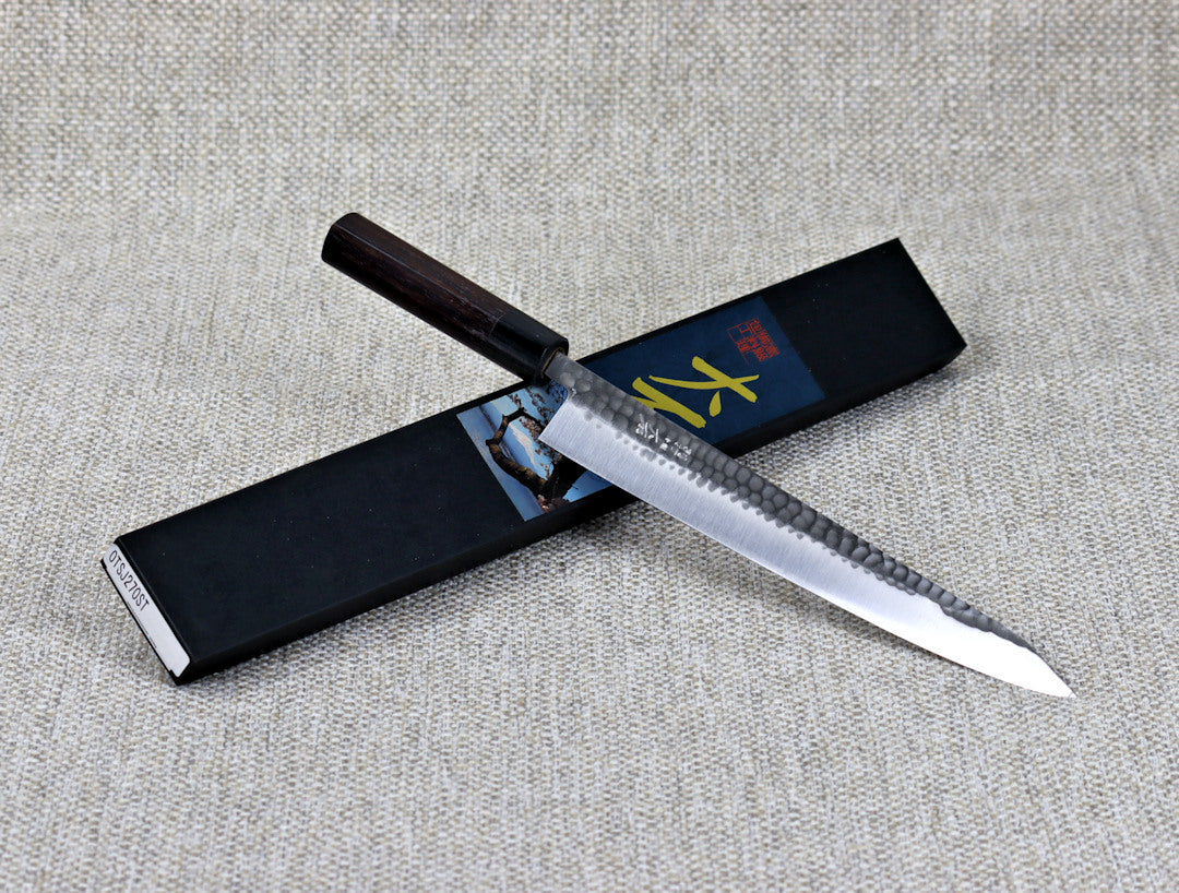 Ohishi SLD Migaki/Tsuchime Sujihiki (Slicer) Japanese Kitchen Knife resting on an angle across its packaging