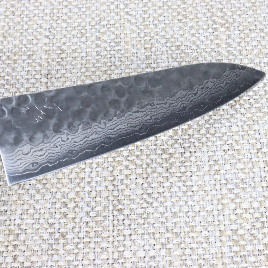 Ohishi Damascus Gyuto (Chef /Cook) hand made Japanese Kitchen knife with Tsuchime （hammered) finish on the top half of the blade and Suminagahsi (floating Ink) Damascus finish at the bottom of the blade. This is a close up of the blade which is slightly raised from the left to show the Kanji style engraving at the handle end of the blade. 
